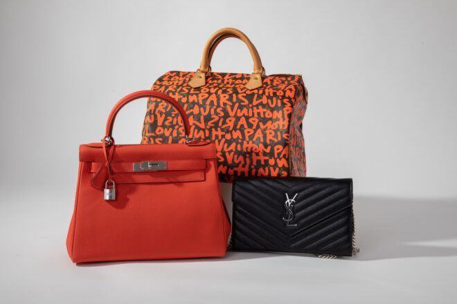 Three handbags: a large orange and black one in the back, an all black clutch in the front and a dark orange next to the black one