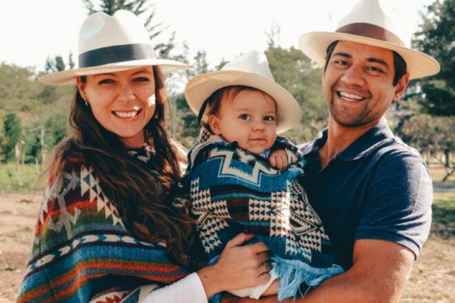 Three people standing within a forest background. From left to right: a woman wearing a hat, a small child wrapped in a colorful blue and black blanket and a man holding the child wearing a matching hat as the woman