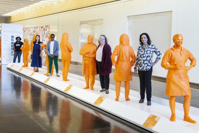 Women standing beside sculptures of themselves that were 3D print in an orange color