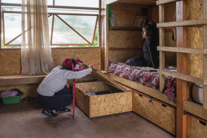 A person sitting on a plywood bunkbed with a person on the floor looks into storage below the bed.