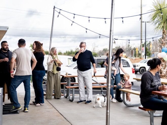 People gathered in the patio space at the front of Green Laundry Lounge. Some are seated at the picnic tables and others are standing near the entrance