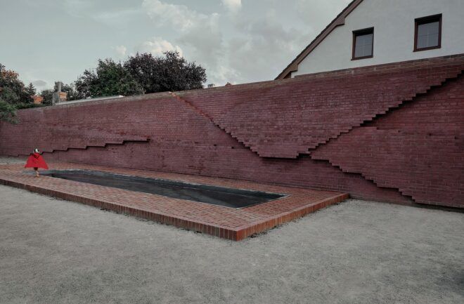 A reddish brown wall with a rectangular pool of water in front of it and a little kid wearing a red top playing beside it