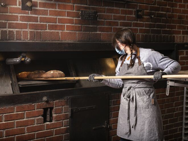A women wearing a blue mask pulls bread out of a bread oven on a wooden paddle
