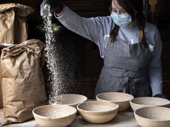 A woman wearing a blue mask sprinkles flour into six bowls