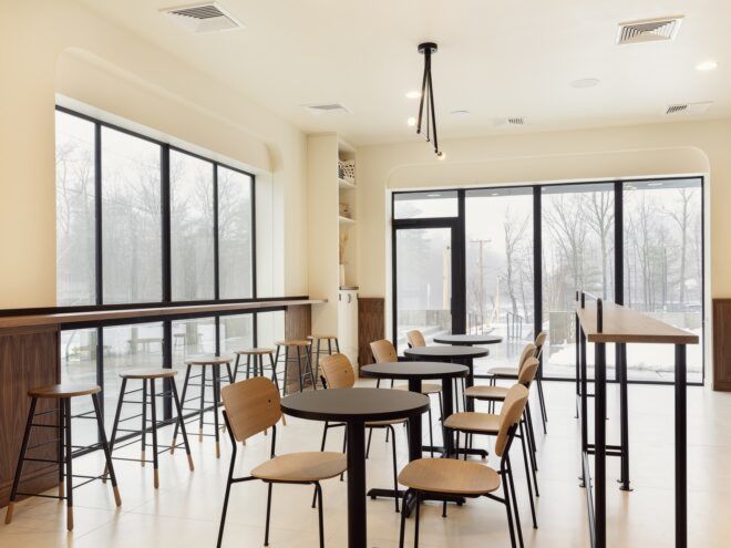 An interior seating area has tables along a window and multiple chairs and tables within the center of the room