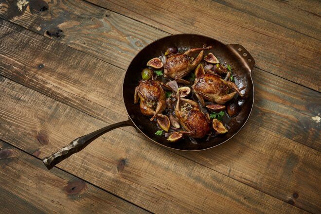 A cast iron pan filled with chicken, figs and herbs sits on dark wood flooring