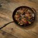 A cast iron pan filled with chicken, figs and herbs sits on dark wood flooring