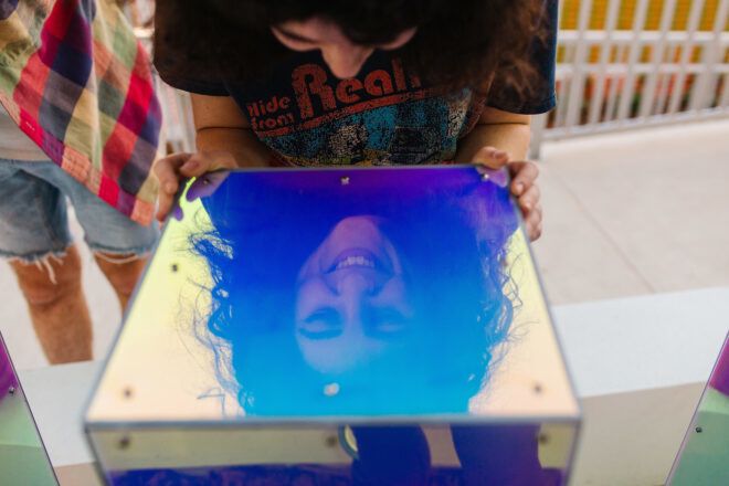 A person holding a reflective, holographic top of the art installation and smiling into their reflection