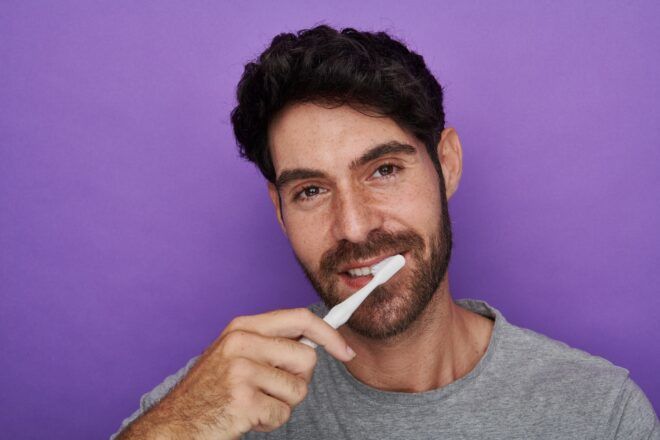 A white male with a beard and black hair has a white Reswirl toothbrush on his teeth standing behind a purple background
