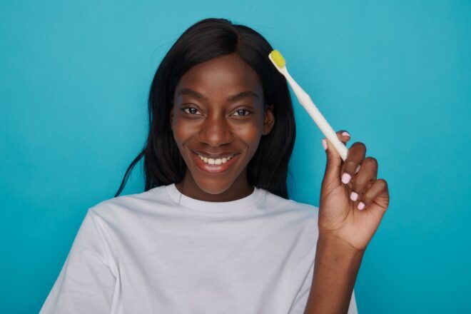 A Black woman holds the white handle of a yellow bristle toothbrush. She wears a white tee shirt and stands in front of a blue background