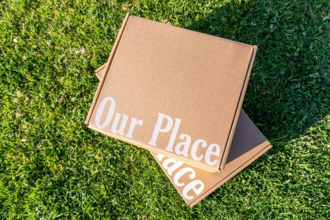 Two cardboard boxes with the words Our Place printed on it in white font, all sitting on grass