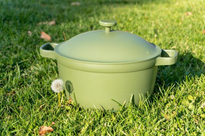 A green pot covered by its lid sits in the grass with a small dandelion planted next to it.