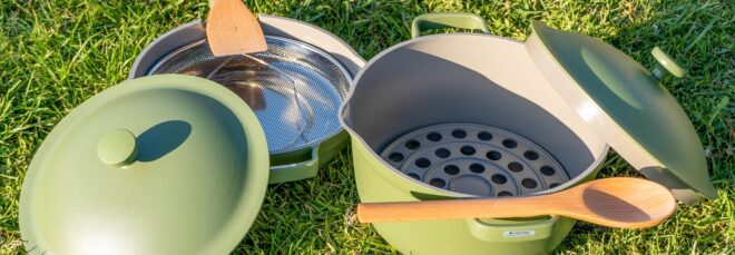 A green pan and pot with their lids leaned against them sits in the grass. In the pan, there is a metal steam rack with a wooden spatula on its handle. Inside the pot, there is a silicone roasting rack with a wooden spoon resting on its handle.