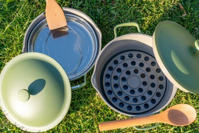 A green pan and pot with their lids leaned against them sits in the grass. In the pan, there is a metal steam rack with a wooden spatula on its handle. Inside the pot, there is a silicone roasting rack with a wooden spoon resting on its handle.