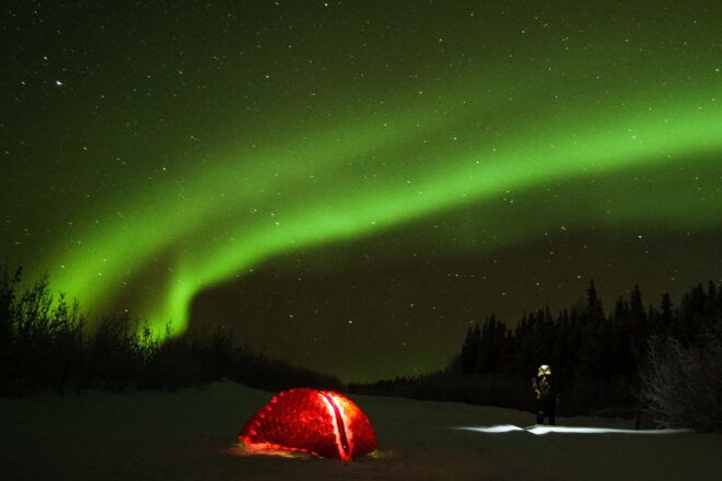 A tent-like shelter lit up in red. The night sky is lit up with green aurora borealis