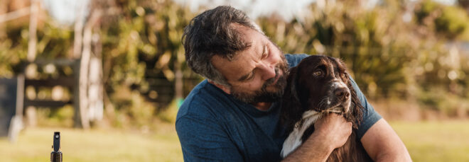 A man in a dark blue shirt hugs a brown and white dog. He is looking at the dog