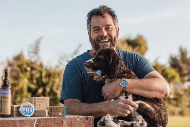 A man looks into the camera wearing a dark blue t-shirt hugs a brown and white dog in his arms with WashBar products set on a table besides him