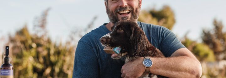 A man looks into the camera wearing a dark blue t-shirt hugs a brown and white dog in his arms with WashBar products set on a table besides him