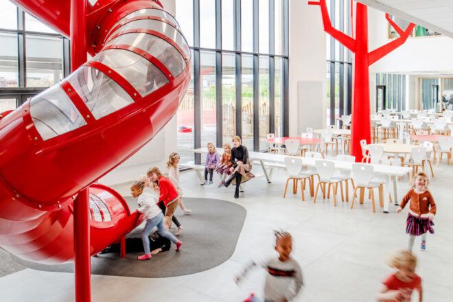 Red and clear slide in a large white and red interior cafeteria space. Lots of children have congregated near the bottom of the slide with some children and a teacher seated nearby