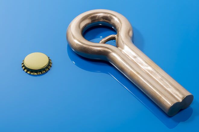 A copper color bottle opener sits besides a bottle cap on a blue paper