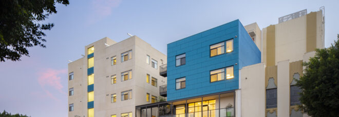 Evening view of a modern, white and blue mid-rise courtyard building facing a street with moving cars