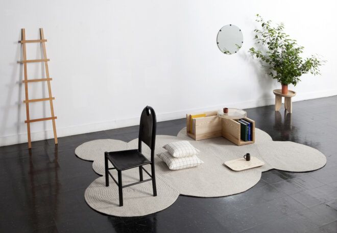 A black floor and white wall area filled with an assortment of furniture: A ladder leans against the wall, a rug, a black chair, a stack of white pillows, a wooden storage unit, a mirror against the white wall and a planter
