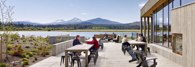 People sitting outdoors on stools that overlook a lake and mountain