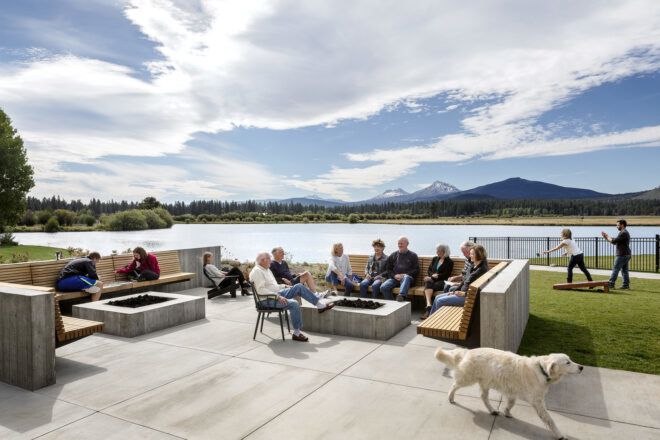 A group of people sit outdoors with an outdoor fire pit in front of them and a lake scenery behind them