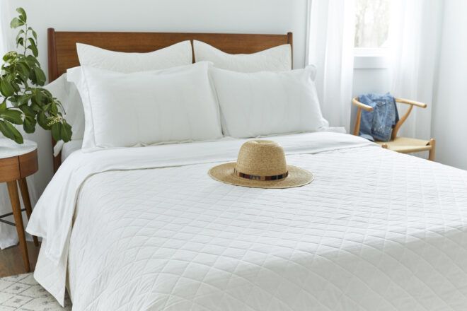A white bed with four white pillows and white covers with a tan wide-brimmed hat sitting on it