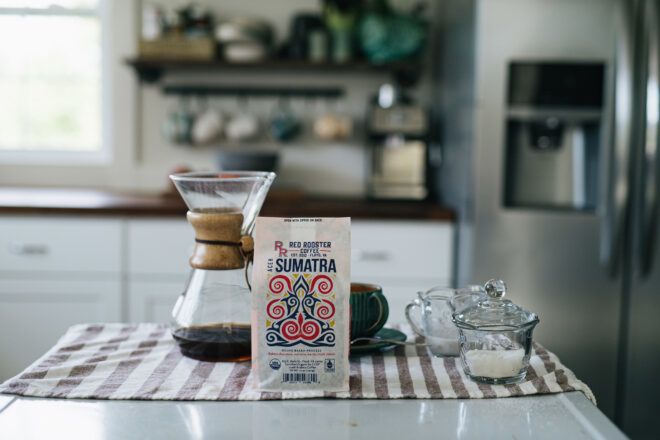 A kitchen scene. At the table is a Red Rooster Coffee bag with a coffee pour over set