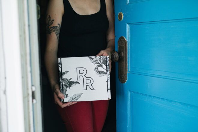 A person leaning against a blue door holding a white box with the initials RR on it