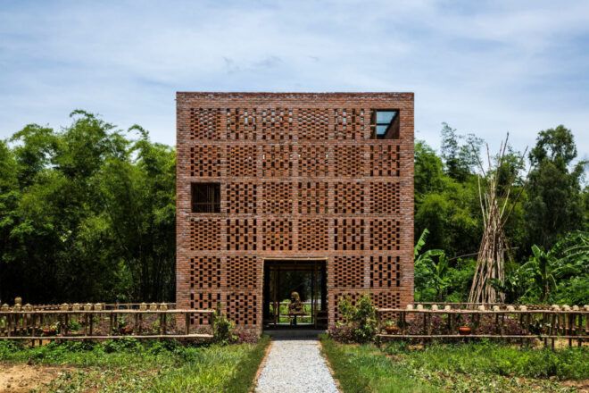 Terracotta brick building in front of a tropical forest. The building facade bricks create a perforated pattern. The building is surrounded by a short bamboo fence