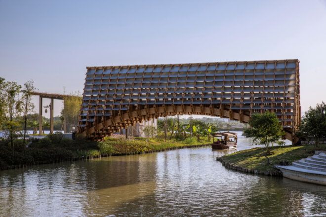 Timber bridge with an arched bottom floating over a waterway. A small boat with many people can be seen about to pass under the bridge
