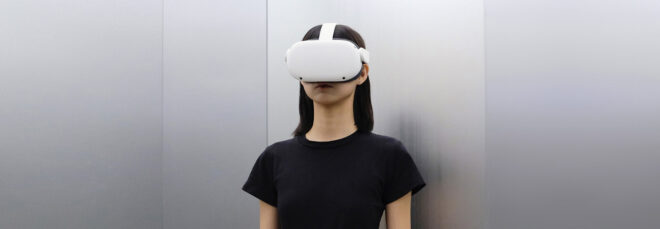 A person wearing a black t-shirt wears a white VR system that covers half their face stands in a metal room