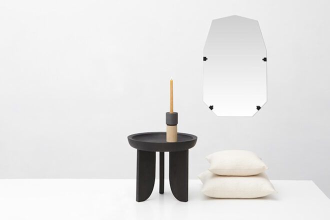 A mirror hangs on a white wall and on the white floor is a brown table with a wooden furniture on it and a stack of white pillows beside it