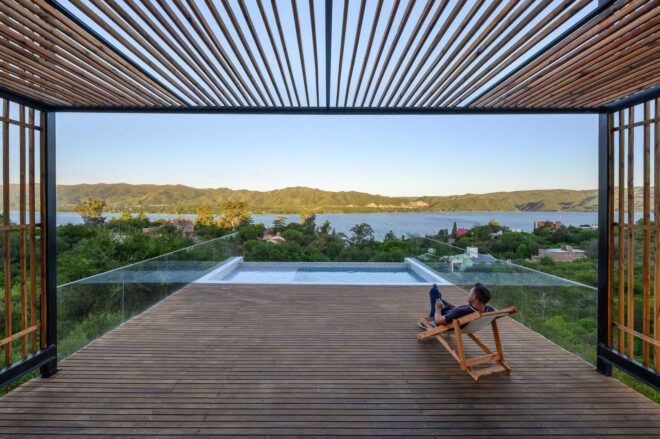 A person reclines on a wooden chair overlooking a pool and river