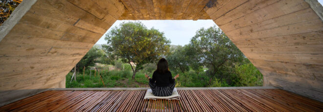 A person sits and mediates underneath a wooden awning overlooking a forest