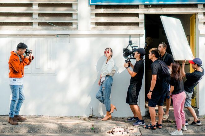 A person on the left, filming a person on the right surrounded by film crew.