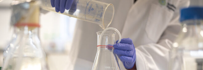 A person holding a cylindrical tube pouring a fluid into another glass beaker