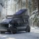 A Canoo electrical vehicle with a camper on the roof sitting in a snowy forest