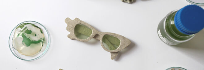 Glasses made out of bacteria sits in the middle of a glass container and a glass bottle with a green liquid