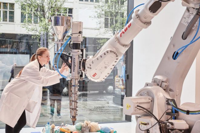 A person wearing a white lab coat is touching a robot that is creating objects out of recycled plastic
