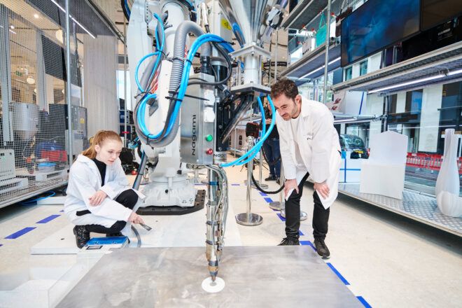 Two people bend down to look at a robot creating something on a platform
