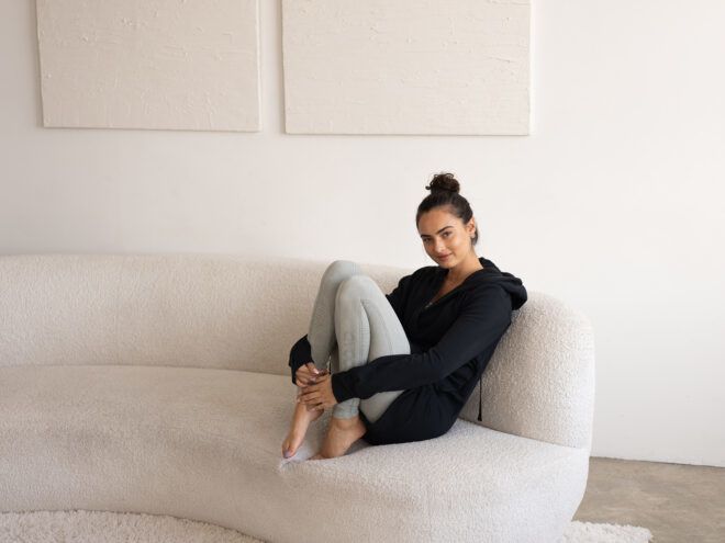 A woman with her legs tucked to her chest wears a black sweater with a grey leggings on a white sofa