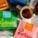 Two coffee bags, one green and one pink, laying beside a cup of coffee with a hand pouring a coffee container