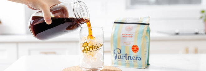 A hand pouring a cup of coffee into a cup filled with ice