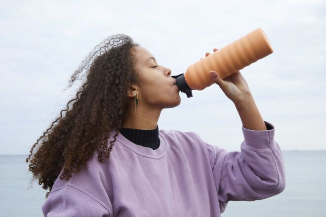 A woman wearing a purple sweater drinks from an orange corrugated bottle