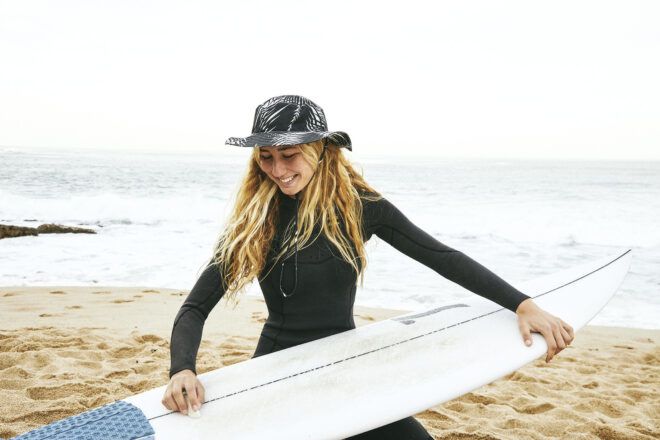 A woman wearing a leaf print black hat and a black wet-suit put wax on a surfboard