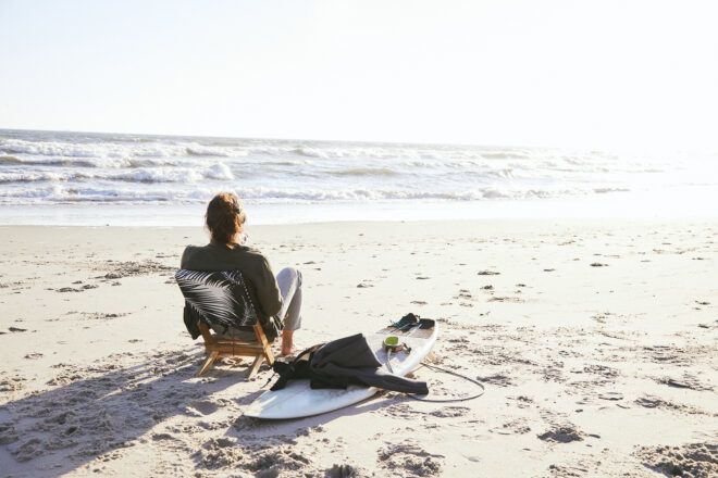 A man sitting on a chair beside an ocean on the sand. A surfboard rests beside him