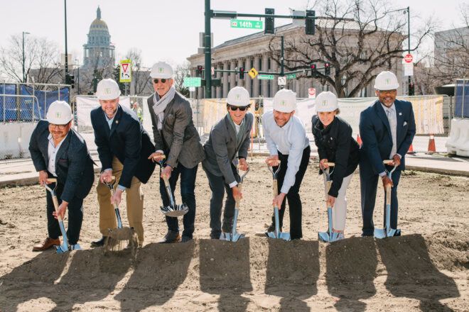 Seven men wearing suits with a white hardhat are bent over their shovels stuck into the dirt